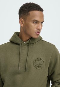 Olive green hoodie made of textured fabric, featuring a circular embroidered logo "NAVIGATE" at the chest and drawstrings on the hood.