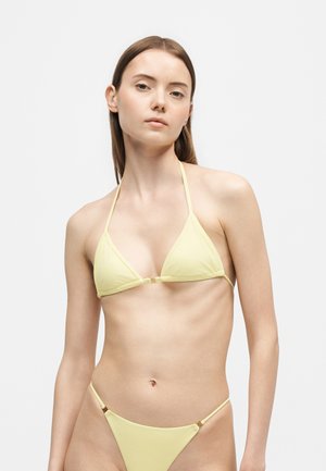 Jeune femme aux cheveux bruns lisses portant un haut de bikini triangle jaune pâle et un bas assorti sur un fond clair uni.