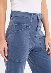 Pantalon utilitaire bleu en mélange de coton avec plusieurs poches plaquées, une taille haute et une finition texturée. Dispose d'une fermeture avec des éléments métalliques.