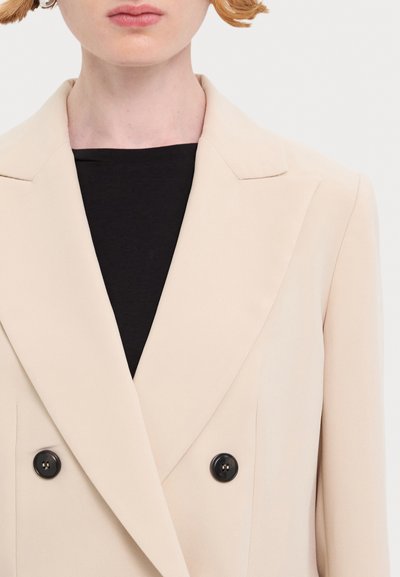Blazer beige à double boutonnage avec des revers élégants, deux boutons noirs et une texture de tissu lisse, superposé sur un haut noir.