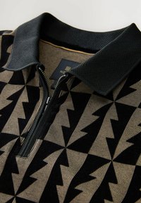 Polo cu închidere cu fermoar, guler negru și un model geometric cu zig-zag în negru și bej. Textură moale cu un design distinct.