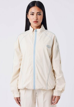 Giacca leggera color crema con accenti azzurri chiari, chiusura zip completa, colletto alto e polsini elasticizzati. Texture liscia e vestibilità rilassata.