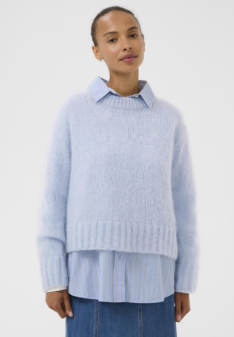 Pull en tricot léger bleu ciel, moelleux, avec ourlet côtelé ; superposé sur une chemise à col rayée bleu clair et blanc ; coupe décontractée et ample.