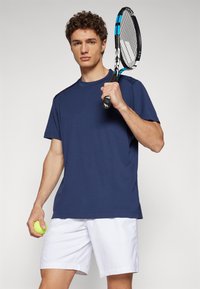 Marineblaues Sport-T-Shirt mit kurzen Ärmeln und rundem Halsausschnitt; kombiniert mit weißen Shorts. Hält einen gelben Tennisball und einen schwarzen Tennisschläger.