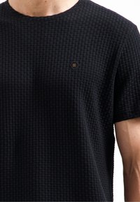 Zwarte texture t-shirt met een geweven patroon, ronde hals en een klein hexagonaal logo in goud op de linkerkant van de borst.