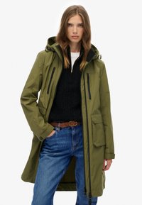 Mujer vistiendo un abrigo con capucha en color verde oliva, suéter negro con cremallera, cinturón trenzado marrón y jeans azules, contra un fondo blanco.