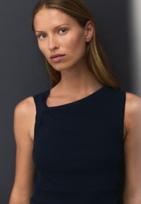 Top sin mangas color azul marino con un escote asimétrico, que presenta un pequeño nudo de tela en el hombro izquierdo. Textura suave.