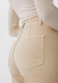 Pantalons taille haute beiges avec une texture lisse, dotés de poches arrière et d'une fermeture éclair dissimulée sur le côté.