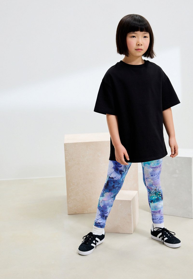 Schwarzes, übergroßes T-Shirt, bunte Leggings mit abstraktem Muster in Blau- und Lilatönen sowie schwarze Sneaker mit weißen Sohlen.