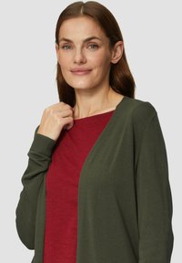 Olivgrüne Strickjacke mit offenem Vorderteil, langen Ärmeln und gerippten Bündchen, getragen über einem roten Shirt. Glatte Textur, lässiges Design.