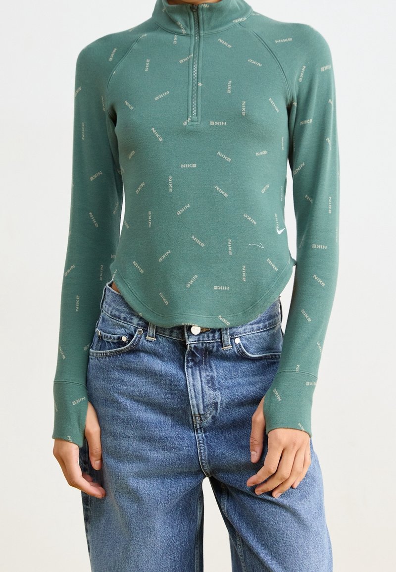 Haut à manches longues couleur sarcelle avec des logos Nike partout, demi-fermeture éclair au col, associé à un jean en denim bleu taille haute.