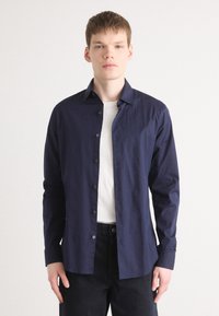 Cămașă bleumarin cu mânecă lungă, dintr-un material ușor, cu guler standard, placket frontal și butoane vizibile. Purtați-o peste un tricou alb.