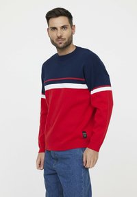 Pull en tricot rouge et marine avec des rayures blanches sur la poitrine, col rond et poignets côtelés. Coupe décontractée avec une étiquette cousue sur le côté.
