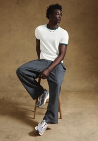 Biała koszulka z zielonym wykończeniem, szare spodnie w prążki oraz szare trampki. Model siedzi na drewnianym stołku na tle neutralnego tła.