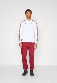 Balta garām piedurknēm polo krekls ar sarkanām svītrām un logo, kombinēts ar sarkaniem sporta biksēm un melniem sporta apaviem. Minimālistisks dizains ar klasisku apkakli.