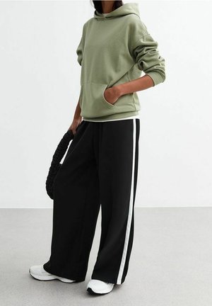 Person trägt salbeigrünen Kapuzenpullover, schwarze breitbeinige Hose mit weißen Seitenstreifen, weiße Turnschuhe und hält eine schwarze Tasche mit Struktur.