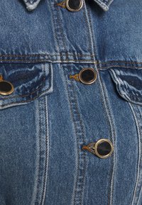 Jeansjacka i blå nyans med guldfärgade knappar, två bröstfickor och kontrasterande sömmar längs skarvarna. Tyget har en mjuk textur.