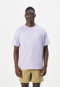 Lys lilla bomull t-skjorte med rund hals, korte ermer og en liten hvit trekantet logo på brystet. Brukt med beige shorts.
