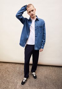 Denim shirt in blauw met knoopsluiting, lichtroze binnenlaag, gecombineerd met zwarte broek en sneakers met witte veters en accenten.