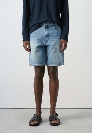 Persona con shorts vaqueros azul claro, camiseta de manga larga oscura y sandalias negras, de pie frente a un fondo liso.