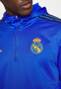 Gros plan sur une personne portant une veste de sport zippée bleue avec le logo jaune et orange du Real Madrid sur la poitrine et des rayures noires sur les manches.