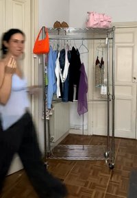 Donna che cammina accanto a un porta abiti in metallo che tiene vari top, una borsa arancione, una borsa da viaggio rosa e scarpe con tacchi alti in una stanza con pavimento in legno.