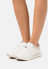 Fötter i vita och beige låga sneakers med snören och sidodragkedjor, stående på en vit yta.