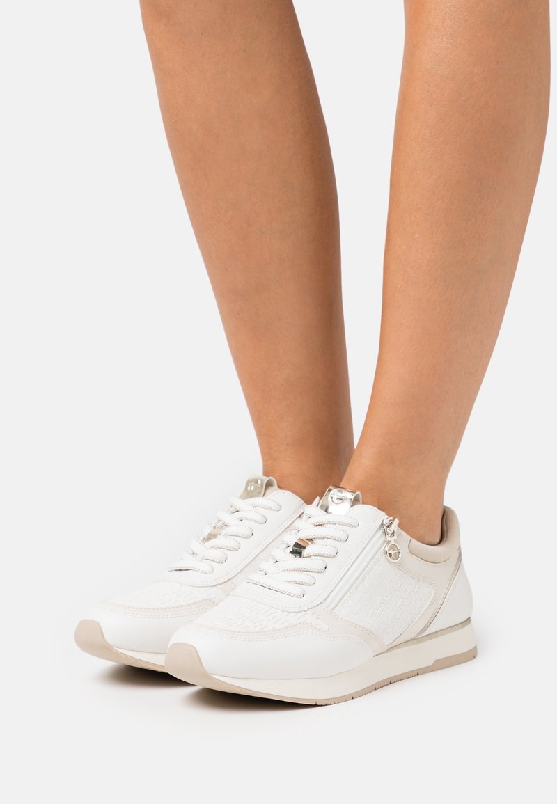 Fötter i vita och beige låga sneakers med snören och sidodragkedjor, stående på en vit yta.