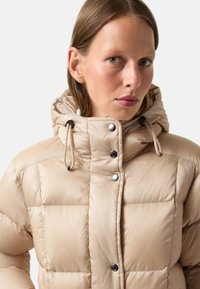 Beige Pufferjacke mit Kapuze, mit einem gesteppten Design, schwarzen Druckknöpfen und verstellbaren Kordeln für eine enge Passform.
