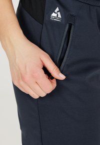 Marinblå WX-TECH-byxor har en slät yta och en sidoficka med dragkedja. En hand som håller i dragkedjehänget framhäver designens detaljer.