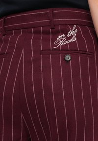 Gros plan sur un pantalon bordeaux avec de fines rayures verticales blanches, un bouton noir sur la poche arrière et un texte brodé blanc indiquant "on the Rocks."