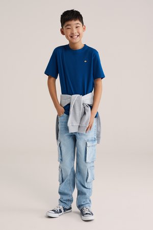 Uśmiechnięty chłopiec w niebieskiej koszulce, jasnoniebieskich spodniach typu cargo, czarnych trampkach i szarej bluzie związanej wokół talii, stojący z rękami w kieszeniach.