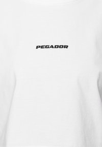 Vit t-shirt med kort ärm som har en svart "PEGADOR"-logotyp över bröstet. Materialet verkar vara slätt, med en klassisk rundhalsad design.