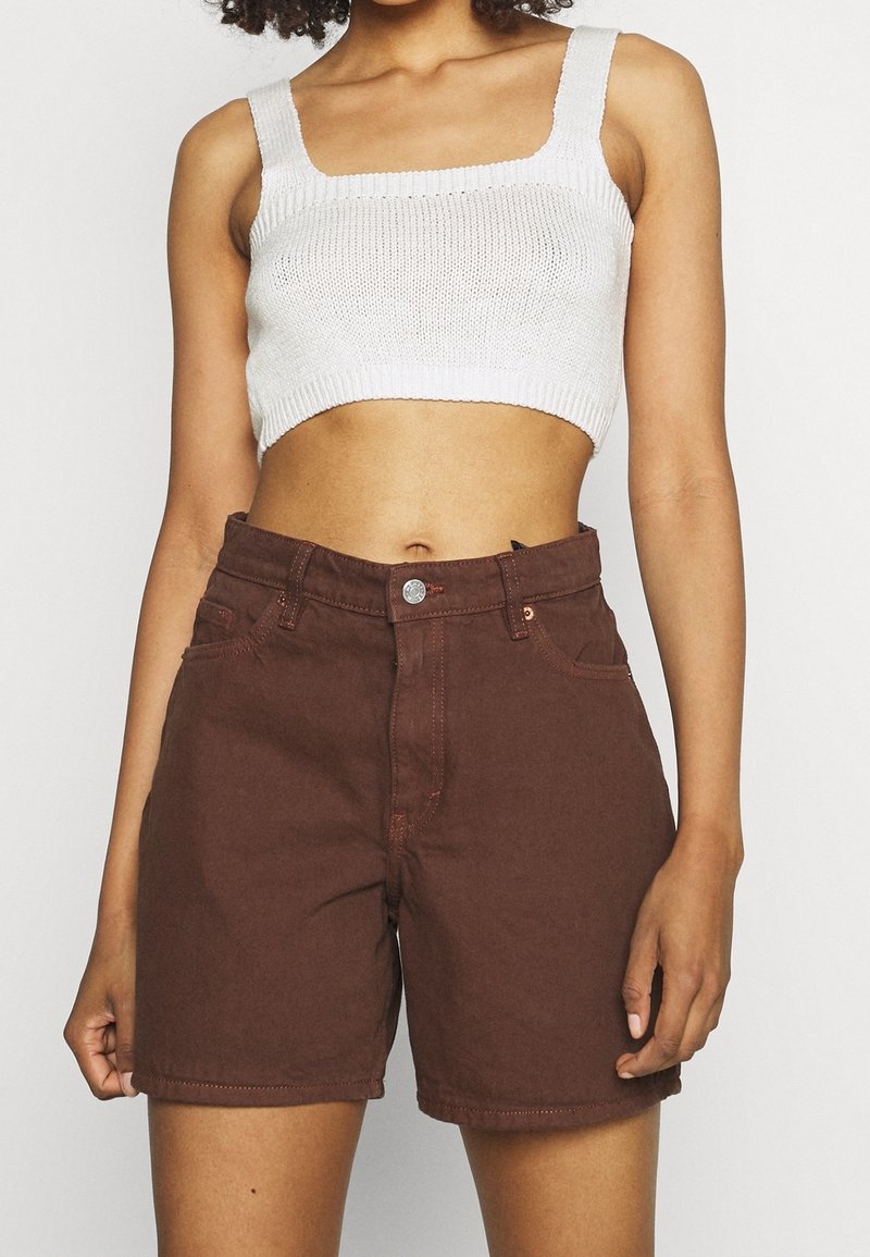 Femme portant un débardeur court en tricot blanc et un short en jean marron taille haute, debout devant un fond uni.