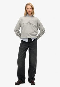 Szara bluza z logo "SUPERDRY", zestawiona z niebieską i białą w paski koszulą pod spodem oraz czarnymi, szerokimi dżinsami. Całość dopełniają brązowe buty.