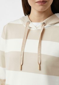 Bluza z kapturem w jasnobeżowe i białe paski z regulowanym kapturem na sznurek. Posiada metalowe przelotki i teksturowaną tkaninę. Emblemat na sznurkach.