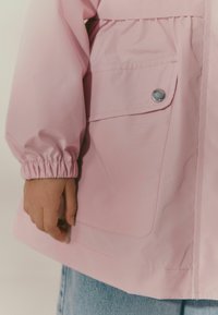 Giacca impermeabile rosa con maniche elasticizzate, tasca con bottone a pressione e vestibilità comoda. Il tessuto appare liscio e leggero.