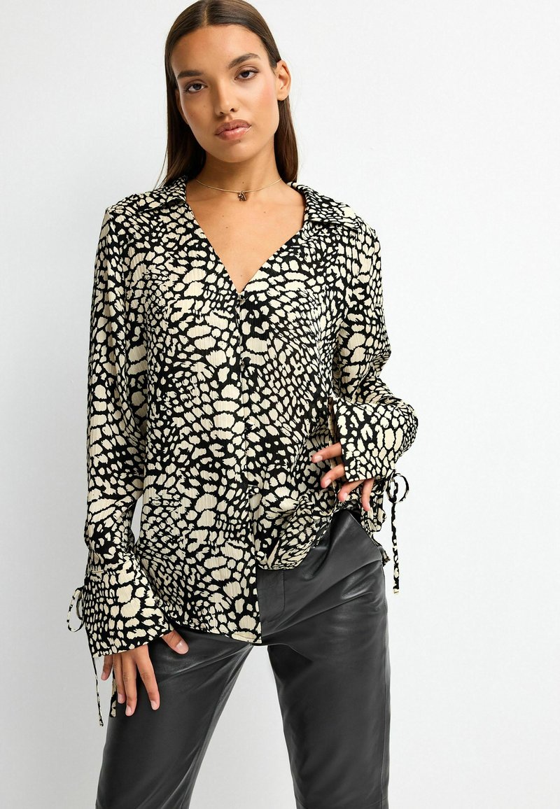 Blouse à motifs noir et crème avec col, patte de boutonnage sur le devant et manches longues. Présente des poignets cintrés et un tissu fluide et léger.