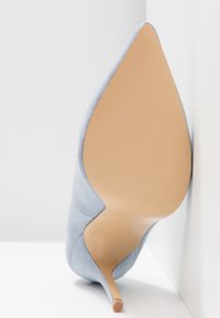 Chaussure à talons hauts en daim bleu clair inclinée pour montrer la semelle beige lisse et le bout pointu sur fond blanc.