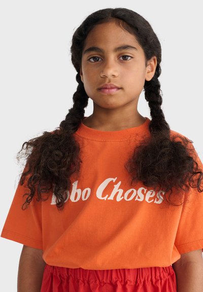 Niña joven con cabello rizado en dos trenzas, con una camiseta naranja con texto blanco y pantalones rojos fruncidos, mirando hacia adelante sobre un fondo claro.