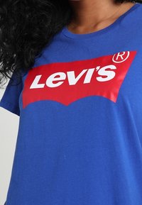 Blå bomullströja med en stor röd Levi's-logotyp och vit text. Rund halsdesign och korta ärmar. Slät textur.