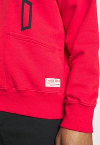 Sudadera roja de Levi's con un bolsillo frontal y puños acanalados. Presenta un diseño gráfico negro y una etiqueta de marca con "Levi's Red" en el bolsillo.