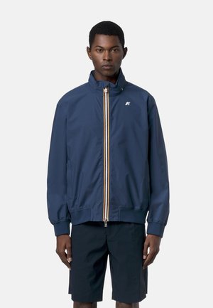 Uomo che indossa una giacca blu navy con zip e collo alto e pantaloncini neri, in piedi su uno sfondo chiaro e semplice.