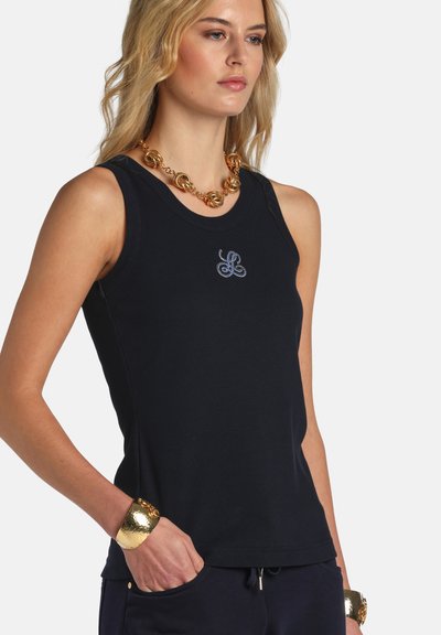 Nő fekete ujjatlan felsőben, kis logóval, aranylánc nyaklánccal és hozzáillő aranykarkötőkkel, kezét zsebében tartva, semleges háttér előtt.