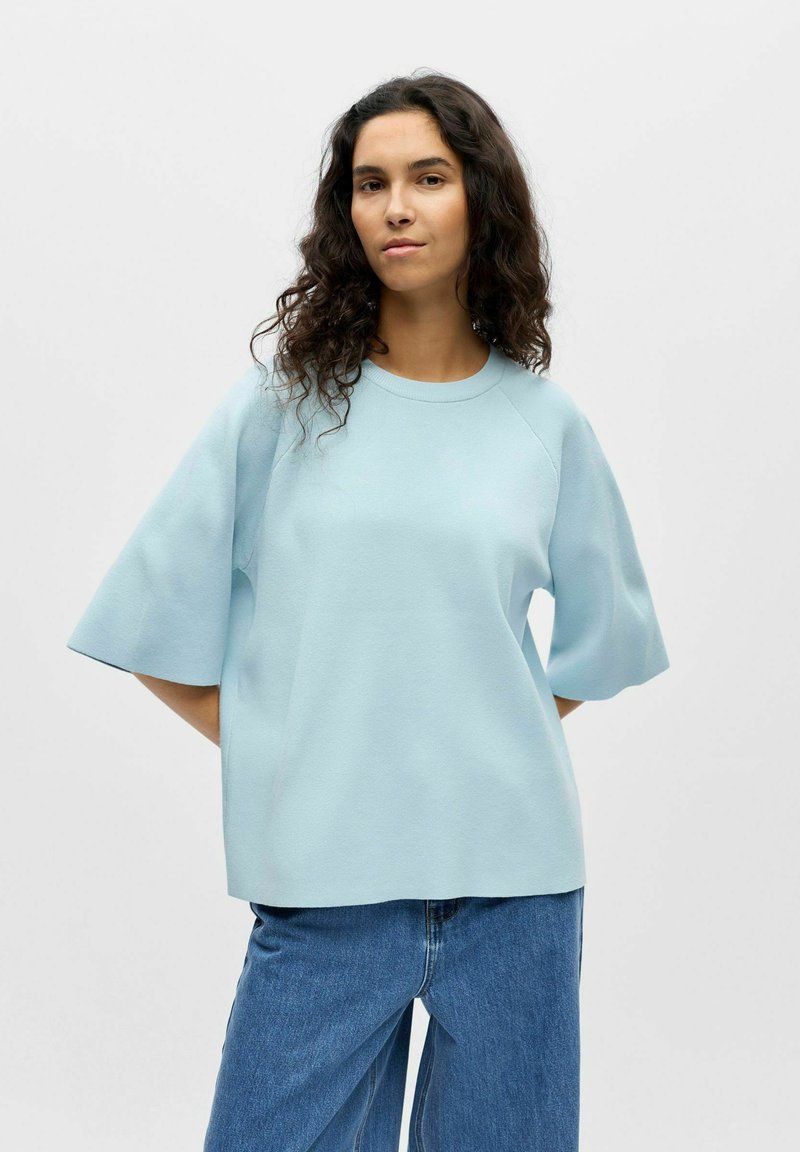 Femme aux cheveux bouclés et foncés portant un haut oversize à manches courtes bleu clair et un jean bleu ample, sur un fond uni.