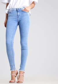 Jeans skinny azzurri chiari in denim, con vita alta, orlo sfilacciato e dettaglio con zip alla caviglia. Indossati con una camicia bianca e tacchi.
