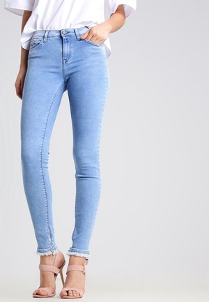 Lichtblauwe skinny jeans van denim, met een hoge taille, rafelige zoom en een ritsdetail bij de enkel. Draagt een witte blouse en hakken.