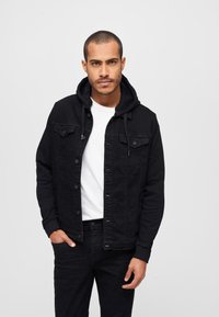 Giacca di denim nera con cappuccio, dotata di due tasche sul petto e chiusura a bottoni. Indossata sopra una t-shirt bianca, abbinata a jeans neri.