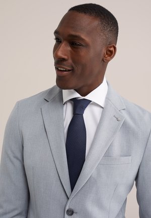 Hombre sonriente, vestido con chaqueta de traje gris claro, camisa blanca y corbata texturizada azul oscuro, mirando hacia la izquierda sobre un fondo claro y liso.