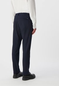 Granatowe spodnie z elastycznym pasem, bocznymi i tylnymi kieszeniami, zwężanymi nogawkami oraz gładką fakturą. Noszone z białą bluzką z długim rękawem.